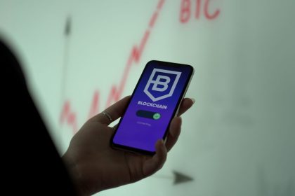 Mão segura um smartphone com um aplicativo de blockchain, enquanto um gráfico financeiro aparece ao fundo.