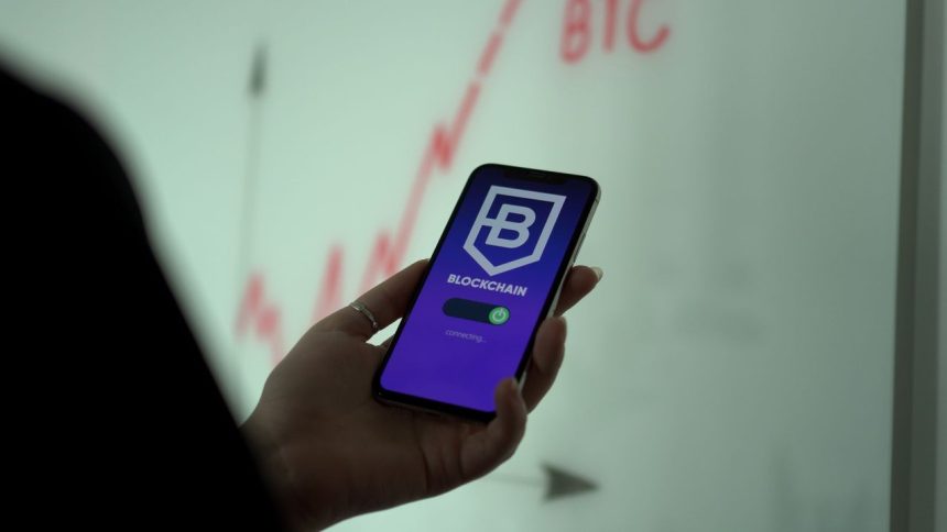 Mão segura um smartphone com um aplicativo de blockchain, enquanto um gráfico financeiro aparece ao fundo.